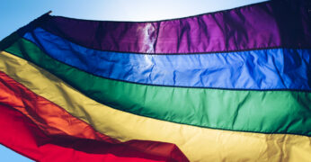 A imagem mostra a bandeira do orgulho LGBTQIAP+ flamulando no céu azul. A foto ilustra o texto sobre população LGBTQIAP+ no mercado de trabalho.