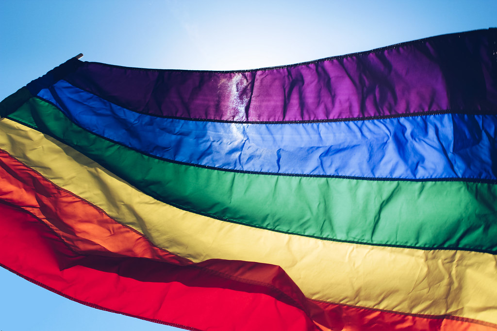 A imagem mostra a bandeira do orgulho LGBTQIAP+ flamulando no céu azul. A foto ilustra o texto sobre população LGBTQIAP+ no mercado de trabalho.