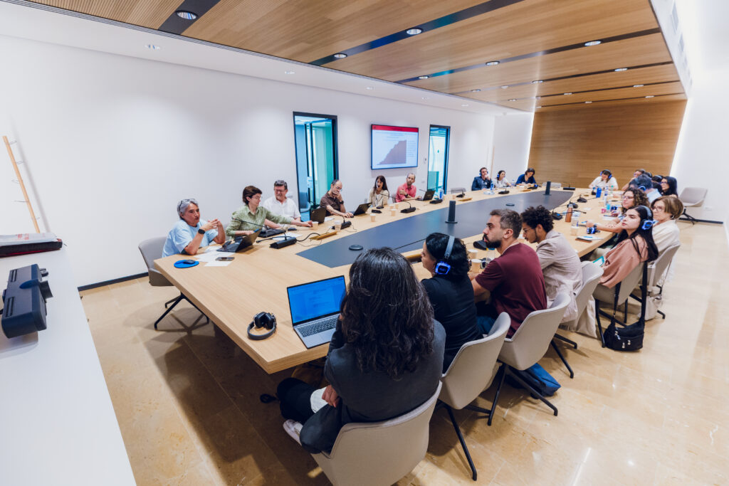 A foto mostra uma reunião de integrantes da comitiva que participou do intercâmbio em Barcelona com representantes do poder público local. As pessoas estão dispostas em uma mesa organizada em formato de "U", de modo que as pessoas de um lado estejam de frente para as do outro. A sala tem uma parede branca, enquanto outra parede e o teto têm revestimento de madeira.