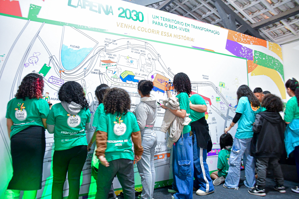 Crianças e adolescentes da Rede Vaga Lume pintam o mapa do Jardim Lapena no banner instalado no palco do Galpão ZL. Elas usam camisetas verdes relativas ao projeto.
