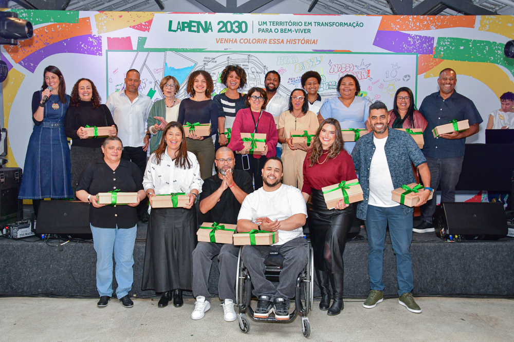 Integrantes da Fundação Tide Setubal estão no palco do Galpão ZL em três fileiras diferentes. Cada uma das pessoas, de gêneros e origens étnico-raciais diversas, seguram uma caixa de madeira com uma fita verde.