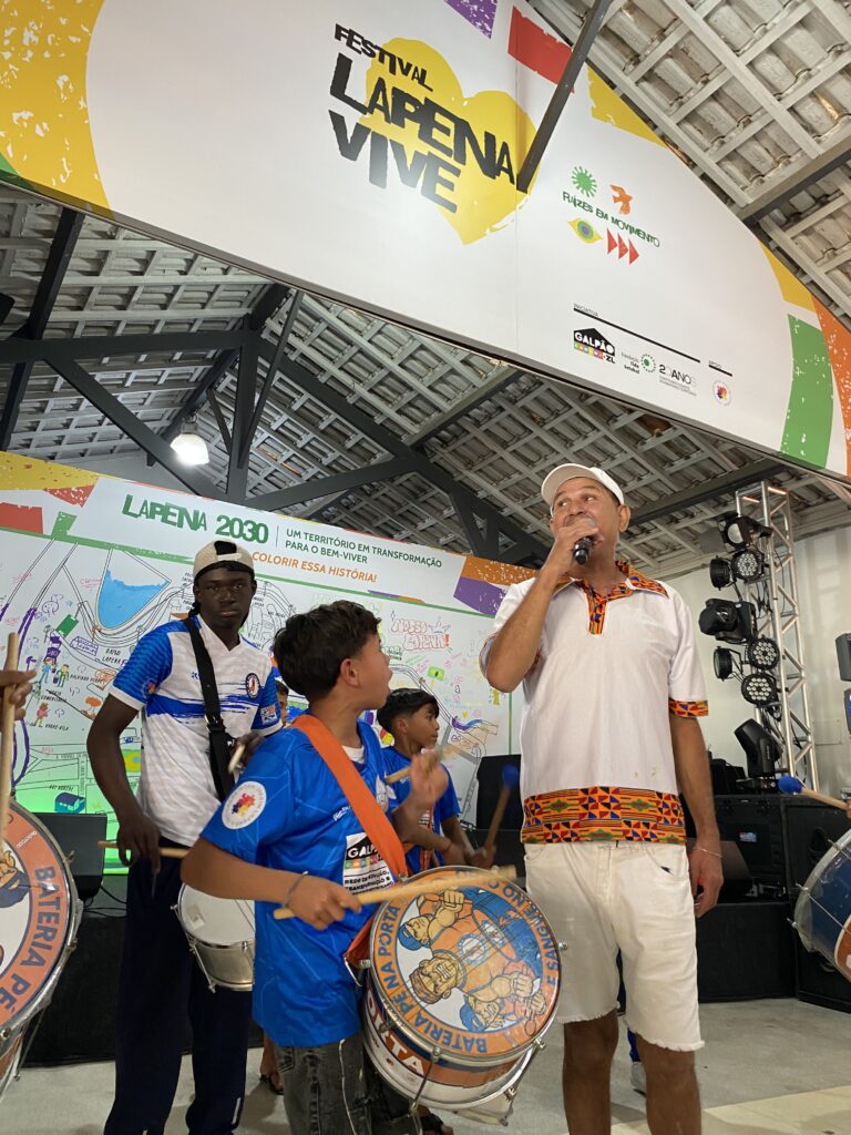 Imagem da Bateria Pé na Porta no palco do Galpão ZL durante o Festival Lapena Vive. Aparecem, no primeiro plano, o intérprete e uma criança tocando um tambor. Demais integrantes aparecem em segundo plano, à frente do painel comemorativo do evento no qual aparece o mapa do Jardim Lapena.