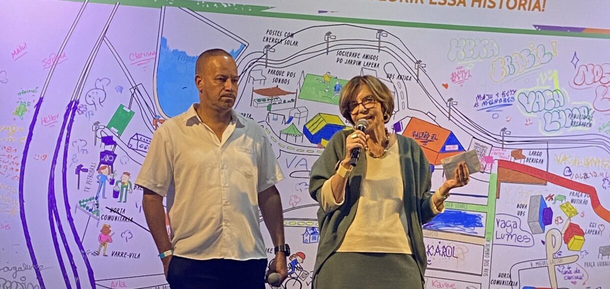 Imagem de Cleiton da Silva (Kaki), presidente da Sociedade Nova Jardim Lapenna (à esquerda), e de Maria Alice Setubal, presidente do Conselho Curador da Fundação Tide Setubal. Maria Alice fala ao usar um microfone e os dois estão no palco do Galpão ZL.