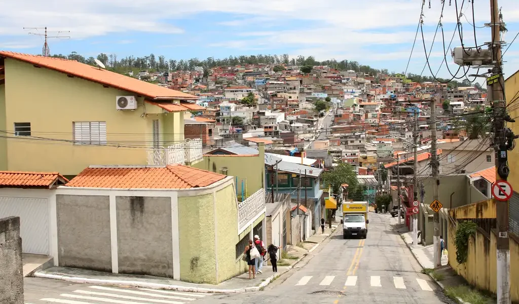 A foto ilustra o horizonte de Perus (São Paulo). Pode-se ver, em primeiro plano, uma casa e a descida de uma rua - há três pessoas andando na calçada à esquerda e um caminhão subindo a mesma via. Aparecem, em segundo plano, diversas casas construídas e uma rua em um morro. A foto ilustra o lançamento da pesquisa 'Espaços Comunitários e Desenvolvimento Territorial nas Periferias de São Paulo'.