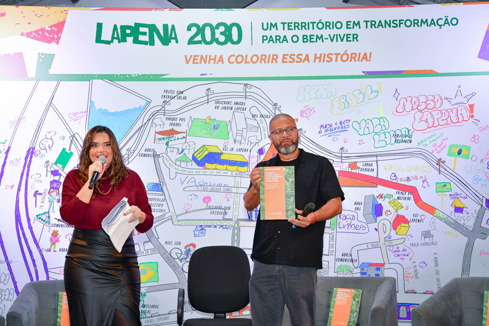 Andrelissa Ruiz e Marcelo Ribeiro Silva estão no palco do Galpão ZL durante o lançamento do livro ‘Galpão ZL, Jardim Lapena: 20 anos fazendo junto com a comunidade’. Andrelissa fala por meio de um microfone e Marcelo segura um exemplar da publicação.