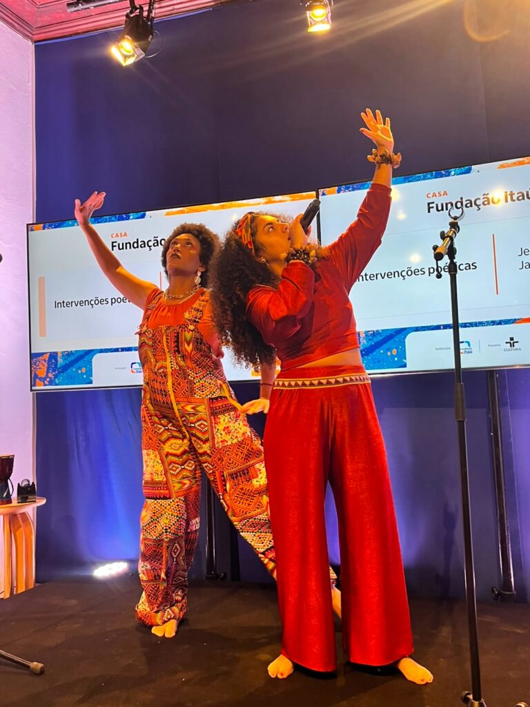A imagem mostra a intervenção Jéssica Marcele (à esquerda, usando jardineira de estampas étnicas e blusa laranja) e Jaque Alves (à direita, com calça e blusa manga longa em tom terroso). Ambas estão no palco da Casa Fundação Itaú + TV Cultura e olham para o alto enquanto estão com os braços erguidos.