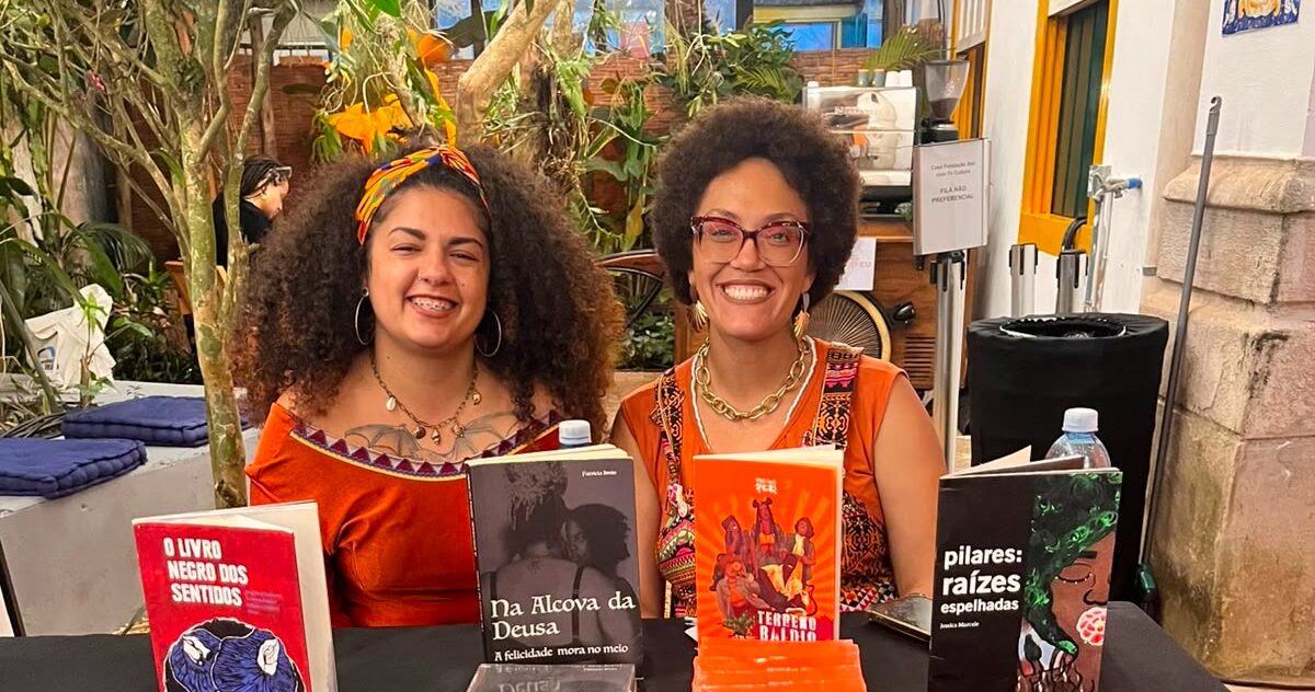 A foto mostra Jaque Alves (à esquerda, com cabelos cacheados) e Jéssica Marcele (à direita, com black power e óculos) em uma mesa da Casa Fundação Itaú + TV Cultura divulgando durante a Flip 2026 os livros dos quais elas participaram e produziram.