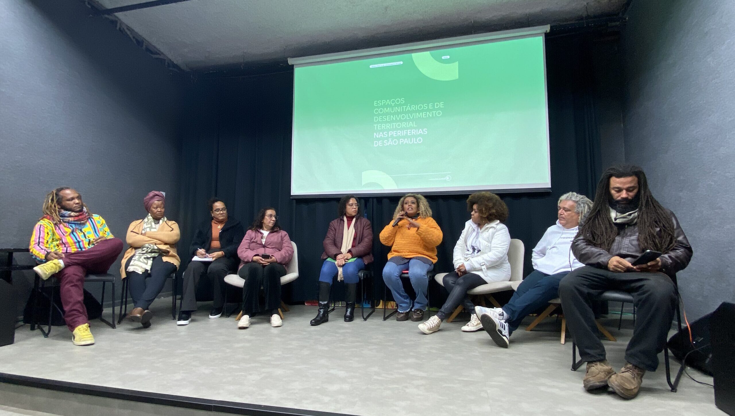 Imagem do evento de lançamento da publicação 'Espaços Comunitários e de Desenvolvimento Territorial nas Periferias de São Paulo'. A foto mostra nove pessoas sentadas em um palco. Há, ao fundo, uma tela com a capa da publicação.