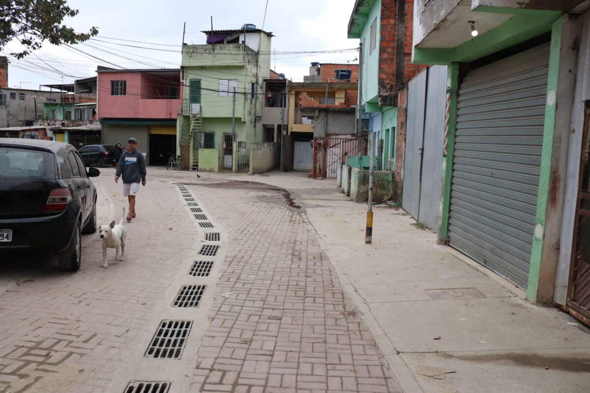 Imagem de uma das ruas do Jardim Lapena após a realização das obras de caminhabilidade. Pode-se ver, à esquerda, um carro, uma pessoa caminhando e um cachorro. Há portões e casas à direita e ao fundo. Ao centro aparecem ruas, calçadas e sistema de escoamento resultantes das intervenções.