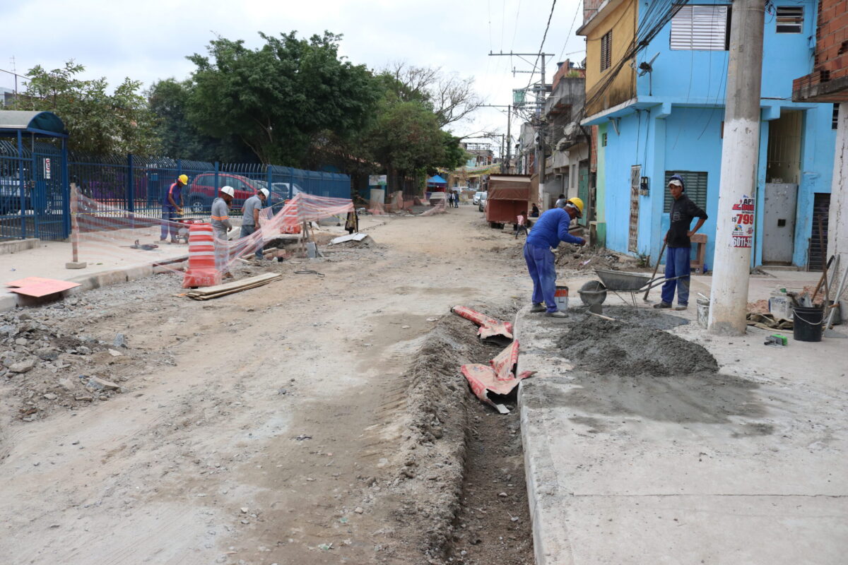 Imagem de obras nas ruas e calçadas do Jardim Lapena. A foto mostra dois profissionais manuseando um carrinho de mão e cimento na calçada. Pode-se ver uma série de casas à direita, uma rua ao centro e árvores no canto superior esquerdo. A foto ilustra a matéria sobre a relação entre comunidade e poder público.