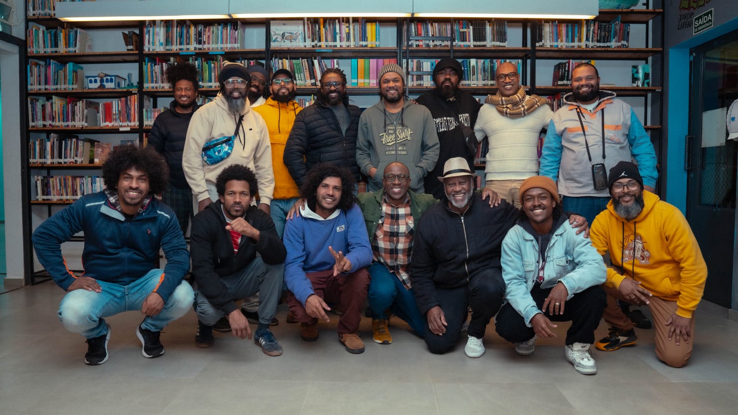 Trata-se de imagem de participantes do Projeto Eu no Singular. Há 16 homens negros (nove em pé e cinco agachados) em frente a uma estante com livros.