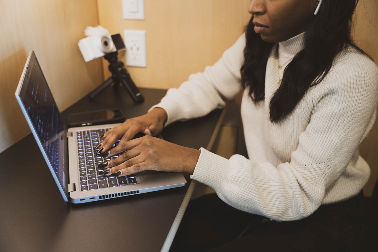Imagem de uma mulher negra usando um notebook. Ela usa uma blusa branca, está com fones sem fio na cor branca e usa uma mesa sobre a qual está também um tripé com uma câmera. A foto ilustra o texto sobre como a aomunicação política e tecnologia podem ser aliadas do bem comum.