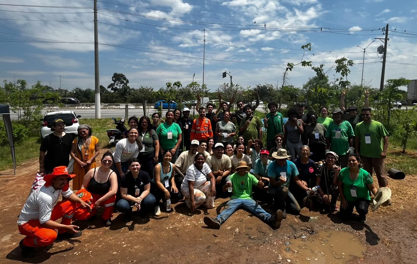 Imagem do grupo de pessoas voluntárias participantes do mutirão para plantio de árvores no Jardim Lapena. Elas estão à frente de uma faixa da avenida Jacu-Pêssego. Há, ao fundo, o céu azul.